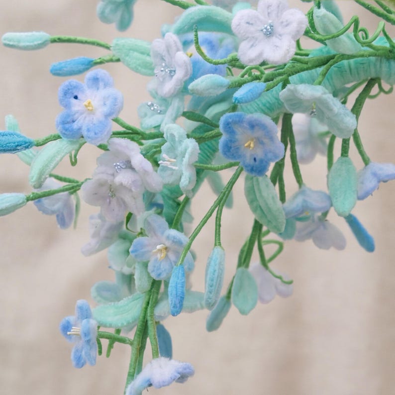 May include: A decorative floral arrangement featuring multiple five-petaled flowers in shades of light blue, white, and soft lavender. The stems and leaves are a vibrant mint green, with small, delicate buds interspersed throughout the branch.