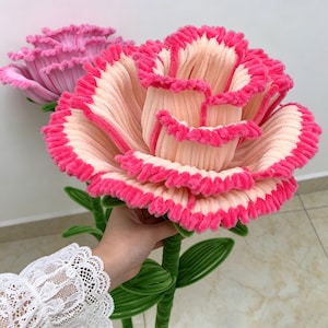 May include: Two large, handcrafted artificial flowers made from textured chenille pipe cleaners. The primary flower features pale peach petals with vibrant hot pink ruffled edges. A second, fully pink flower is visible in the background.
