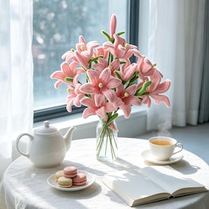 May include: A bouquet of plush pink lily flowers with green stems arranged in a clear glass vase. The arrangement features multiple open blooms and one closed bud, displayed on a round table alongside a white teapot, a cup of tea, and macarons.