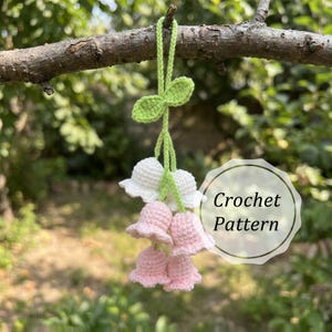 May include: A handmade crochet floral decoration featuring white and pink bell-shaped flowers, green leaves, and a green hanging cord. The text "Crochet Pattern" is visible on the image.