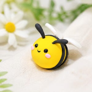 May include: A small, round, yellow and black bee figurine made of smooth material. The bee features two black antennae, two white wings, two small black eyes, and two pink circular cheeks on its face, with a simple black line for a mouth.