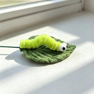 Könnte beinhalten: Eine leuchtend hellgrüne, flauschige Raupe aus Pfeifenreinigern mit zwei großen weißen Wackelaugen mit schwarzen Pupillen, die auf einem strukturierten, dunkelgrünen Stoffblatt mit einem dünnen grünen Drahtstiel an der Seite ruht.