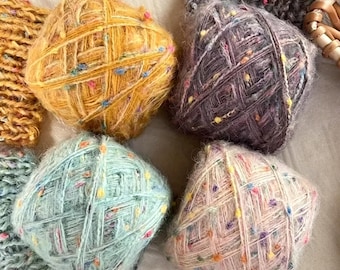 May include: Four balls of fuzzy yarn in yellow, dark gray, light blue, and pale pink, each featuring a textured surface with small, multicolored flecks throughout. The yarn appears soft and slightly fibrous, arranged in a square-like formation.