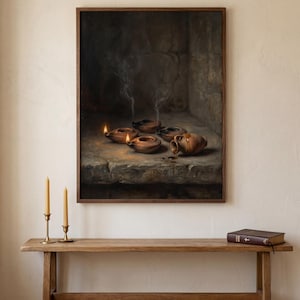 May include: A framed wall art print depicting a dark, stone-textured interior scene with several small, lit clay oil lamps emitting thin wisps of smoke. A small, rounded clay pitcher sits beside the lamps on a rough, uneven stone surface.