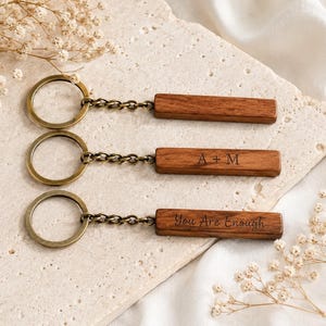 Può includere: Tre portachiavi in legno con anelli e catene color ottone. Un portachiavi ha il testo inciso "You Are Enough", e un altro ha le iniziali incise "A + M". I portachiavi sono marroni.
