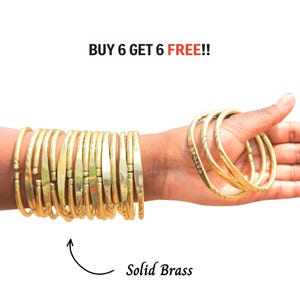 May include: A collection of gold-colored metal bangles with a smooth, polished finish. The text BUY 6 GET 6 FREE!! is visible at the top, and the text Solid Brass is visible at the bottom with an arrow pointing toward the bangles.