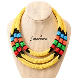May include: A multi-strand beaded necklace featuring three layers of tubular, seed-bead ropes. The design incorporates yellow, black, blue, orange, green, and red beads arranged in geometric patterns. The text LuxeArena is printed in the center.