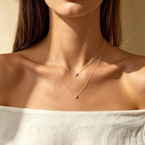 Pode incluir: Dois colares delicados com pequenas pedras redondas e transparentes em engastes de metal. Um colar é feito de metal prateado, enquanto o outro é de metal dourado. Ambas as correntes são finas e apresentam uma única pedra central.