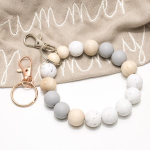 Könnte beinhalten: Ein Schlüsselanhänger-Armband mit einem roségoldenen Verschluss und einem silbernen Verschluss. Das Armband hat Holzperlen in verschiedenen Farben, darunter weiß mit Sprenkeln, hellgrau und Naturholztöne. Das Wort "summer" ist auf den beigen Stoff gestickt.
