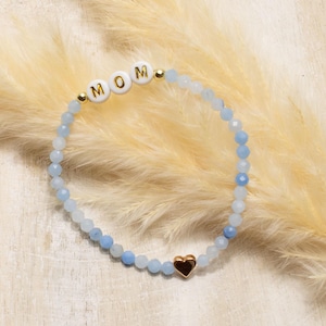 Könnte beinhalten: Ein Perlenarmband mit hellblauen facettierten Perlen, drei weißen runden Perlen mit goldener Aufschrift MOM, zwei kleinen goldenen Zwischenperlen und einem einzelnen herzförmigen Anhänger in Goldoptik am unteren Ende des Armbands.