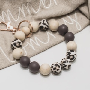 Könnte beinhalten: Ein Schlüsselanhänger-Armband mit Holzperlen in Creme, Dunkelgrau und Leopardenmuster. Das Armband hat einen goldfarbenen Verschluss und Ring und liegt auf einem beigen Stoff.