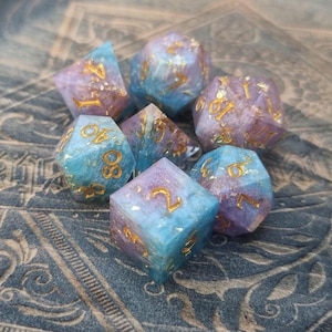 May include: A set of seven polyhedral dice featuring a swirling blend of light blue and soft purple resin with embedded gold-coloured flakes. Each die has engraved numbers filled with gold paint, including a d20, d12, d10, d8, d6, d4, and a percentile die.