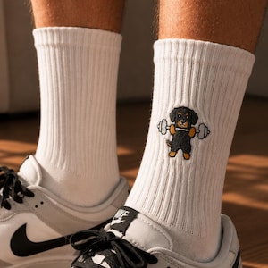 Op de afbeelding: Witte geribbelde enkelsokken met een geborduurde afbeelding van een zwart-bruine hond die een grijze halter optilt. De sokken worden gedragen in wit-grijze lage sneakers met zwarte veters en een zwart logo aan de zijkant van de schoen.
