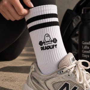 Può includere: Calze bianche a girocollo con due strisce nere sulla parte superiore, caratterizzate da un ricamo di un fantasma che solleva un bilanciere con la scritta DEADLIFT sottostante. Indossate con una scarpa sportiva grigia e bianca con logo N nero.