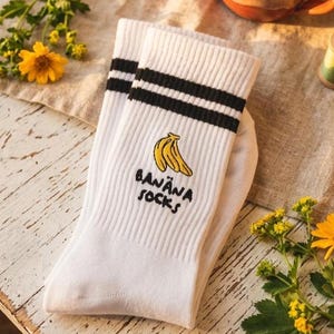 Könnte beinhalten: Weiße Socken mit schwarzen Streifen und einer Bananen-Grafik. Die Socken haben die Worte "BANANA SOCKS" unterhalb der Banane gestickt. Die Socken sind auf einer weißen Holzoberfläche gefaltet, mit einer Tasse und Garnrollen im Hintergrund.