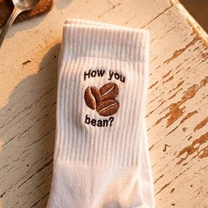 Può includere: Calzini bianchi con la frase ricamata "How you bean?" e grafica di chicchi di caffè. I calzini sono piegati e poggiano su una superficie di legno bianco usurato. Una tazza di caffè e un cucchiaio sono visibili sullo sfondo.