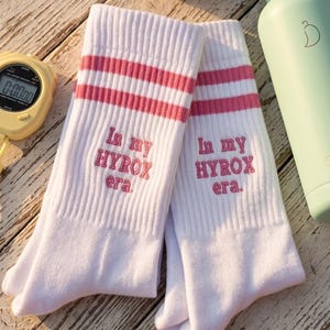 May include: White athletic socks with pink stripes and the embroidered text "In my HYROX era." A rolled-up lavender towel, a light green water bottle, and a stopwatch are also visible on a weathered wooden surface.