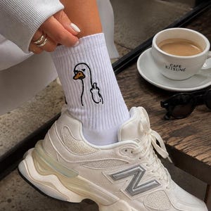 May include: White ribbed crew socks featuring an embroidered graphic of a goose making a rude hand gesture, paired with white athletic trainers that have silver accents and a thick, layered sole design with a prominent N logo on the side.