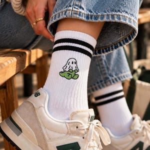Könnte beinhalten: Weiße Crew-Socken mit zwei schwarzen Streifen am oberen Rand und einer kleinen gestickten Grafik eines Geistes, der auf einem grünen Klecks sitzt. Die Socken werden mit cremefarbenen und beigen Sportschuhen mit grünem N-Logo kombiniert.