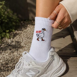 May include: A white ribbed crew sock featuring an embroidered graphic of a goose wearing sunglasses and red high-heeled shoes with the text SLAY in red lettering, paired with a white athletic trainer with mesh and silver accents.