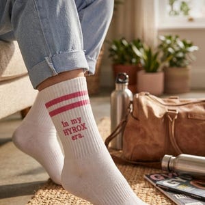 May include: White athletic socks with two pink stripes and the text "In my HYROX era." The socks are ribbed and worn with blue jeans. A gym bag and water bottles are in the background.
