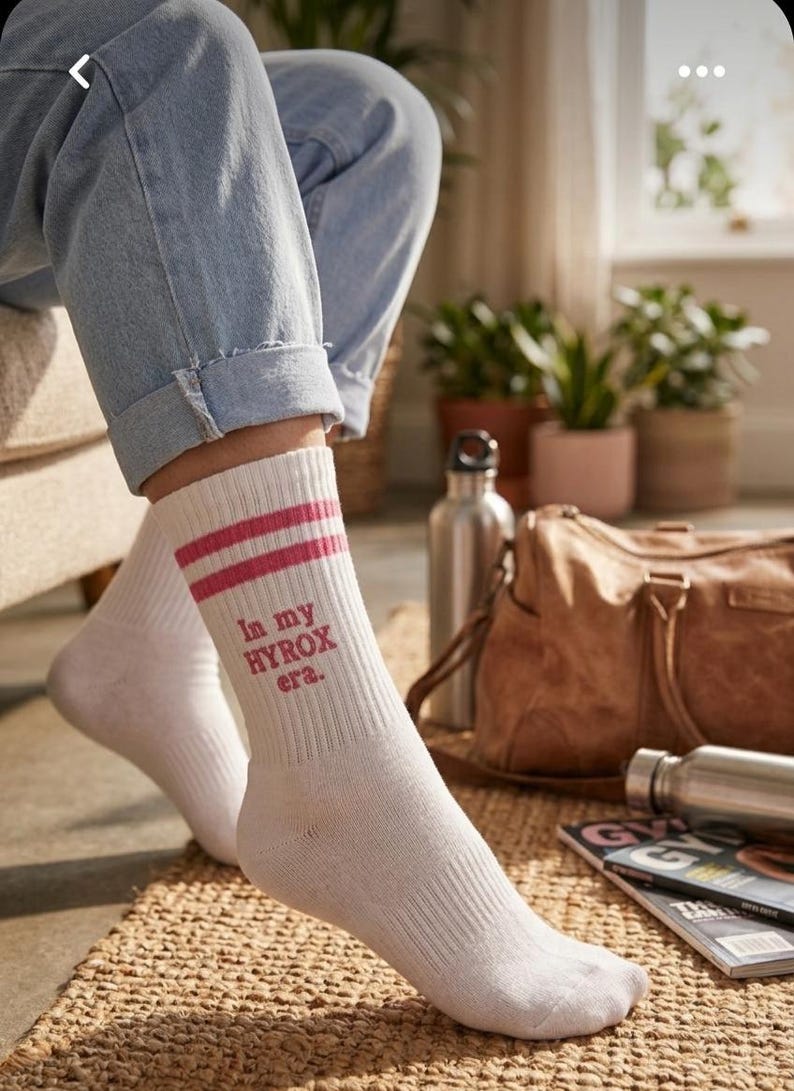 May include: White athletic socks with two pink stripes and the text "In my HYROX era." The socks are ribbed and worn with blue jeans. A gym bag and water bottles are in the background.
