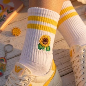 Könnte beinhalten: Weiße Socken mit gelben Streifen und einer Sonnenblumenstickerei. Die Socken werden mit weißen und gelben Turnschuhen getragen. Ein Notizbuch und Accessoires befinden sich im Hintergrund.