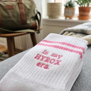 May include: White socks with pink stripes and the embroidered text "In my HYROX era." The socks are folded on a grey surface. A green duffel bag and potted plants are visible in the background.
