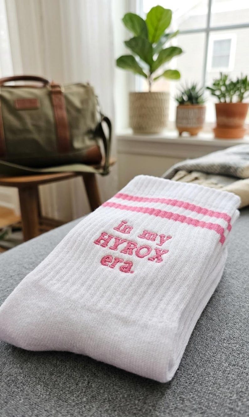 May include: White socks with pink stripes and the embroidered text "In my HYROX era." The socks are folded on a grey surface. A green duffel bag and potted plants are visible in the background.
