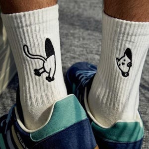 Puede incluir: Calcetines blancos acanalados con ilustraciones de gatos negros de dibujos animados, combinados con zapatillas deportivas azules y turquesas con suelas marrones. Los calcetines muestran un diseño de gato.