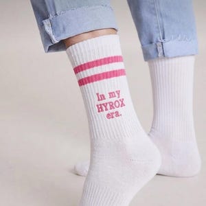 May include: White athletic socks with two pink stripes and the text "In my HYROX era." in pink. The socks are worn with blue jeans.