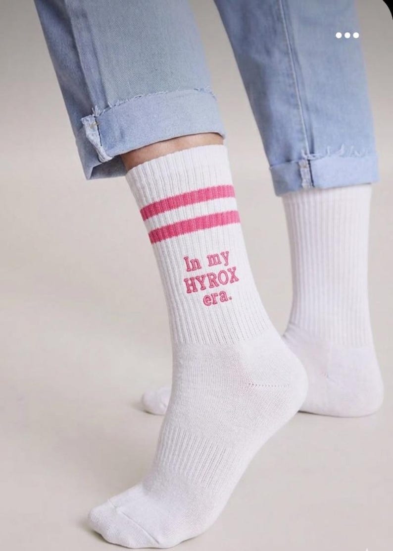 May include: White athletic socks with two pink stripes and the text "In my HYROX era." in pink. The socks are worn with blue jeans.