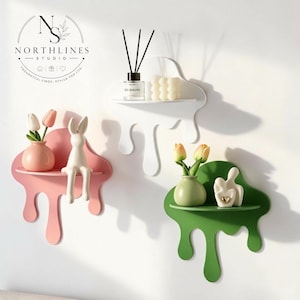 May include: Three wall-mounted shelves shaped like melting liquid in white, pink, and green. The pink shelf holds a small vase with two tulips and a rabbit figurine. The white shelf holds a reed diffuser and a bubble candle. The green shelf holds a vase with two tulips and a small abstract figurine.