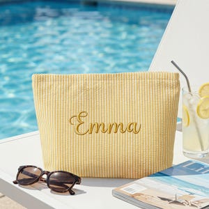 Könnte beinhalten: Eine gelbe Seersucker-Kosmetiktasche mit dem Namen "Emma" in goldener Schrift gestickt. Eine Sonnenbrille mit Schildpattmuster liegt vor der Tasche. Ein erfrischendes Getränk und eine Zeitschrift sind in der Nähe.