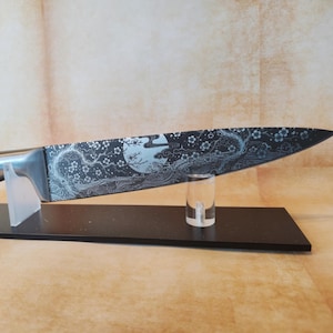 Può includere: Un coltello da chef in acciaio inossidabile con una lama incisa decorativa con un design a luna e floreale. Il coltello poggia su un supporto nero con un supporto in acrilico trasparente. L'impugnatura è argentata.