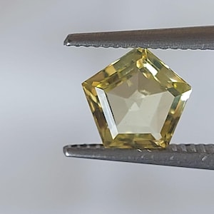 Può includere: Una gemma sfaccettata a forma di pentagono, tenuta da una pinzetta. La gemma ha una tonalità giallo pallido e un design simmetrico con molteplici sfaccettature. La pinzetta è color argento e lo sfondo è di un tono neutro.