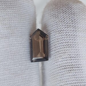 Könnte beinhalten: Ein facettierter, schildförmiger Rauchquarz-Edelstein mit einem durchscheinenden, bräunlich-grauen Farbton. Der Stein hat eine spitze Oberseite und eine flache Basis und zeigt präzise geometrische Schliffe, die das Licht reflektieren.