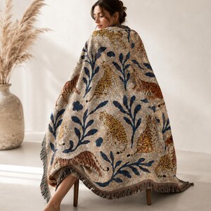 Puede incluir: Una manta beige con un estampado de leopardos y hojas azules. La manta tiene un borde con flecos y está sobre una persona sentada. Un textil para el hogar decorativo para un ambiente acogedor.