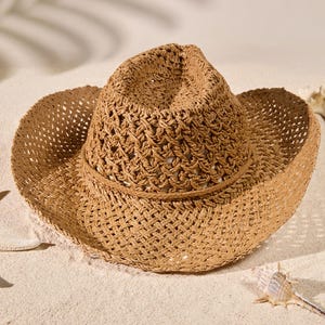 Pode incluir: Um chapéu de cowboy de palha bege com uma estrutura de tecido aberto, uma copa vincada e uma aba curva. O chapéu é acentuado com uma fita simples de palha trançada fina que circunda a base da copa, apresentando um acabamento texturizado.