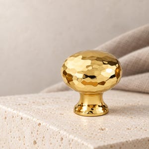 Può includere: Un pomello per mobili color oro con una texture martellata e riflettente. Il pomello presenta una superficie superiore arrotondata e leggermente appiattita e una base liscia e affusolata che si collega al punto di fissaggio, creando un aspetto metallico lucido.