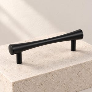 May include: A matte black cabinet pull with a tapered, hourglass-like silhouette. The handle features a smooth, curved central section that widens toward both ends, supported by two cylindrical posts extending downward from the main body.