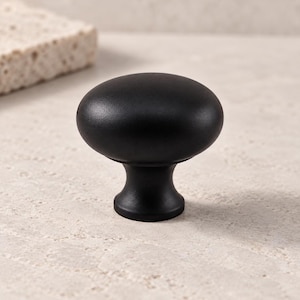 May include: A matte black cabinet knob featuring a smooth, rounded, mushroom-shaped head and a tapered, circular base. The hardware has a solid, uniform dark finish with a clean, curved silhouette designed for attachment to furniture or doors.