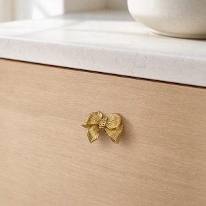 Peut inclure: Un bouton de meuble en métal doré en forme de nœud décoratif avec une surface nervurée et texturée. Le nœud présente une bande centrale avec de petits détails en relief, fixé sur une façade de tiroir en bois clair au grain visible.