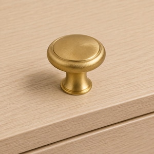 Puede incluir: Un pomo de mueble circular con acabado en oro cepillado. El pomo presenta una superficie superior plana y redonda con un sutil detalle de anillo concéntrico y una base cilíndrica cónica que se conecta a la superficie de montaje.