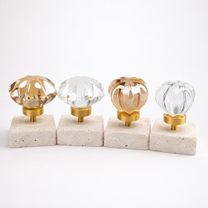 Puede incluir: Cuatro pomos para armarios con cabezas de cristal estriado en forma de calabaza, alternando tonos transparentes y ámbar. Cada pomo está unido a una base metálica de tono latón, montada sobre un bloque de piedra beige cuadrado y poroso.