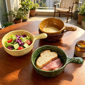 Puede incluir: Tres cuencos de cerámica con asas, cada uno con un acabado moteado. Un cuenco amarillo claro contiene una ensalada de lechuga, pepino y tomate. Un cuenco verde oscuro contiene una rebanada de pan y jamón. Un cuenco amarillo mostaza contiene sopa.