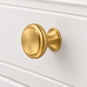 May include: A round cabinet knob with a brushed gold finish and a smooth, circular face. The knob features a tapered base that connects to a flat, circular mounting plate, creating a clean, uniform appearance for furniture hardware.