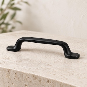 May include: A matte black cabinet handle with a smooth, arched design and rounded ends. The hardware features a solid, uniform dark finish with a curved grip that transitions into flat, oval-shaped mounting points on each side of the handle.
