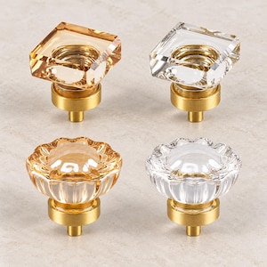May include: Four cabinet knobs featuring faceted glass tops in square and flower shapes. Two knobs have clear glass, and two have amber-colored glass. Each knob is mounted on a circular brushed brass base with a central metal post for attachment.