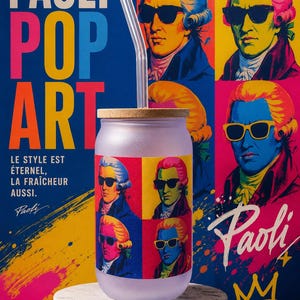 Peut inclure: Un verre givré avec un couvercle en bambou et une paille en verre transparent. Le verre présente une impression pop art colorée d'un personnage historique portant des lunettes de soleil, reprenant le motif de l'affiche PAOLI POP ART en arrière-plan.